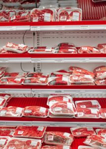 A home cook places sealed raw meat on the lowest refrigerator shelf beside an appliance thermometer.